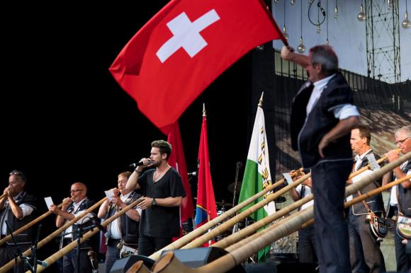 Il cantante Bastian Baker mentre intona l inno svizzero attorniato da suonatori di corni delle alpi e da una bandiera svizzera.