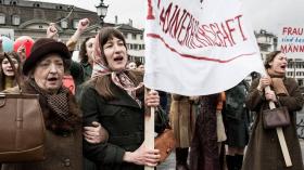 Scena di una manifestazione per il suffragio femminile: delle donne innalzano uno striscione e gridano.