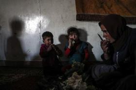 Due bambini con la mamma seduti per terra in un angolo stanno mangiando, mettendo il cibo in bocca con le mani.