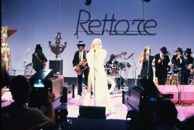 Donatella Rettore vestita completamente di bianco sul palco con musicisti tutti in abito nero. Sul fondo, la scritta Rettore