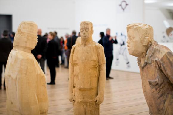 Primo piano di tre sculture che rappresentano individui trisomici. Sullo sfondo, visitatori della mostra.