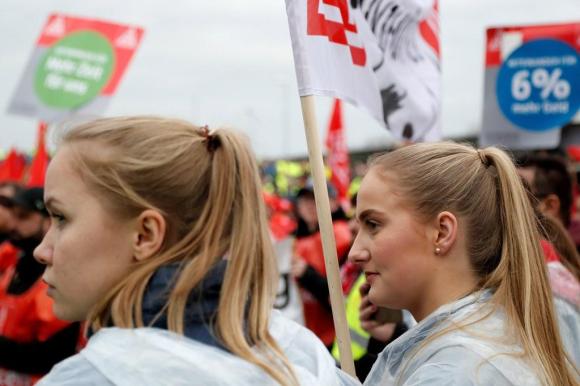 Manifestazione di lavoratori, con due giovani donne in primo piano e cartelloni e bandiere sullo sfondo