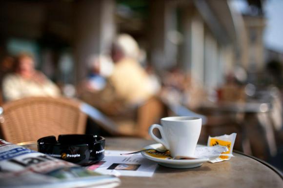 Una tazzina di caffè, una bustina di zucchero e un giornale su un tavolino all aperto. Avventori del bar sfocati.