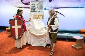 Donna agghindata da Helvetia e uomo vestito da Guglielmo Tell indicano un manifesto a favore di Moneta intera