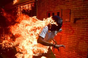 Giovane uomo con maschera antigas e t-shirt corre avvolto dalle fiamme; un muro sullo sfondo