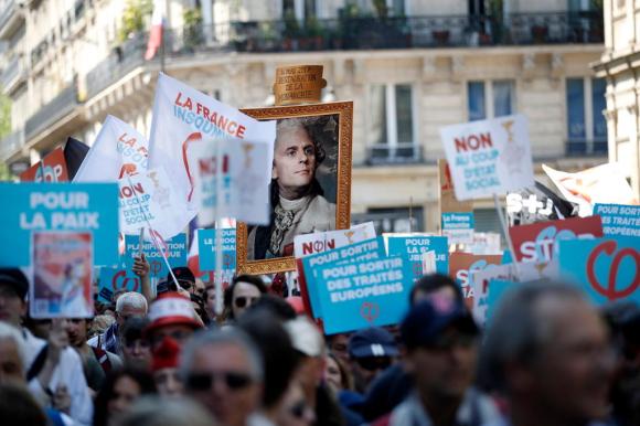 Un ritratto di Macron dipinto come un monarca, a fuoco tra numerosi cartelli con vari slogan portati da manifestanti in strada