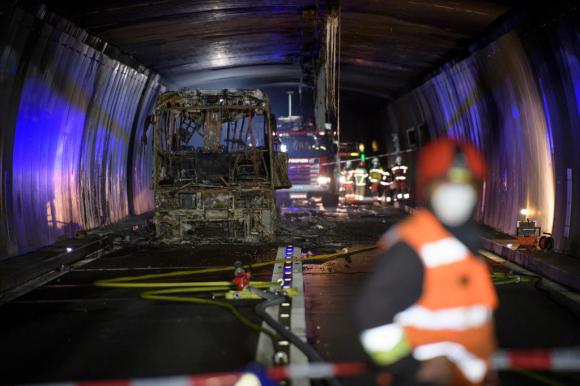 Interno semibuio di un tunnel stradale; sulla sx la carcassa di un autobus; sulla dx pompieri con mantellina catarifrangenti