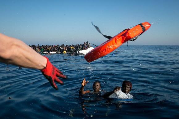 Volontario dell Ong Sea Watch lancia un salvagente a due ragazzi caduti in mare. Sullo sfondo un barcone carico di migranti