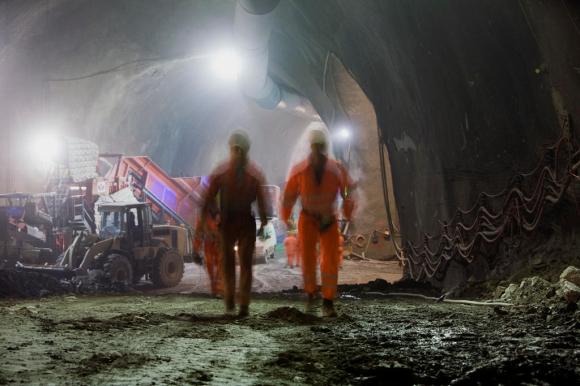 Due operai (sfocati) camminano all interno di un tunnel illuminato da forti torce; macchinari di scavo intorno
