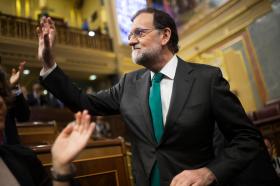 Primo piano di mariano Rajoy che alza la mano in segno di saluto; sullo sfondo, scranni del parlamento sfocati