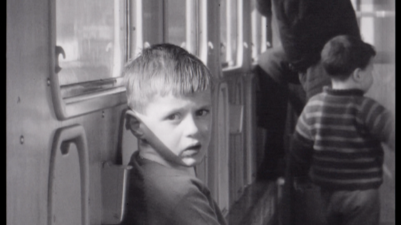Bambino ripreso sul vagone di un treno accanto a un finestrino, guarda in camera; in fondo un altro bimbo di schiena