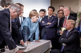 merkel e altri membri del G7 in piedi parlano a Trump seduto su una sedia