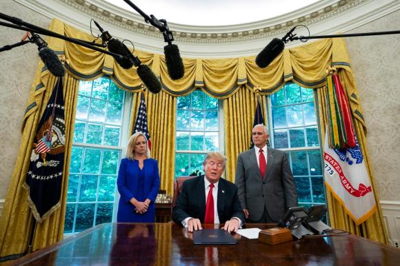 Trump alla scrivania, nel gesto di chiudere una cartelletta contente il decreto firmato; a sx la segretaria Nielsen, a dx Pence