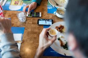 Un tavolo di ristorante, con stoviglie e persone che mangiano: al centro due smartphone che mostrano le ultime notizie