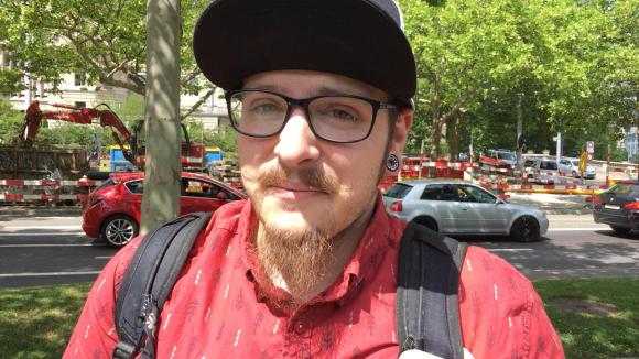 Ein Mann in rotem T-shirt mit Hut und Brille schaut in die Kamera.