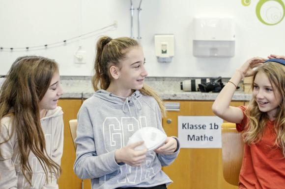 Tre ragazze sorridenti sedute in un aula stanno provando ad indossare la kippa.