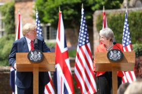 Trump e May ai rispettivi pulpiti si guardano e si sorridono; dietro, 3 bandiere USA e 3 UK alternate