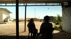 Una donna col velo e un uomo, nascosti dal contrasto con lo sfondo, dove si intravede un campo di prigionia nel deserto