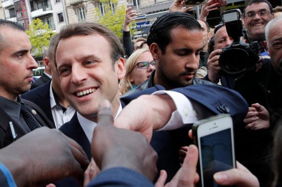Primo piano di Macron; subito dietro di lui, Benalla; circondati dalla folla (mani e telefonini protesi verso il presidente)