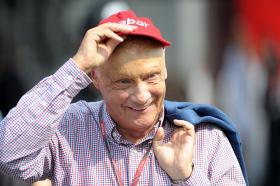 Primo piano di Niki Lauda che sorride e solleva la visiera di un cappellino rosso; indossa camicia a quadretti rossi e blu