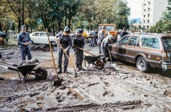 Viale alberato cittadino completamente coperto da fango; uomini in uniforme al lavoro con pale e carriole