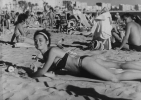 Donna distesa al sole, sulla spiaggia; un uomo dietro di lei la guarda; sul fondo lungomare con palazzi