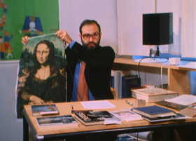 Umberto Eco, nel suo studio, mostra un grembiule con stampata la Gioconda; libri sulla scrivania