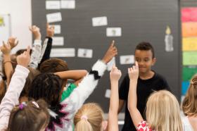 Ein Junge steht vor einer Schulklasse, seine Kameraden und Kameradinnen strecken die Hände in die Luft.