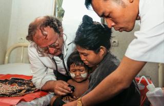 A man with a stethoscope inspects a child, who is held by a woman.