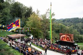 Folla segue una cerimonia officiata dal Dalai Lama in ambiente collinare; sventolano bandiera del Tibet e bandiere di preghiera
