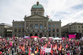 Folla di manifestanti di fronte a palazzo federale con bandiere, cartelli e palloncini di colori rosso, rosa e fucsia