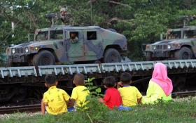 Bambini seduti sul ciglio della strada guardano passare veicoli blindati con militari armati di mitra.