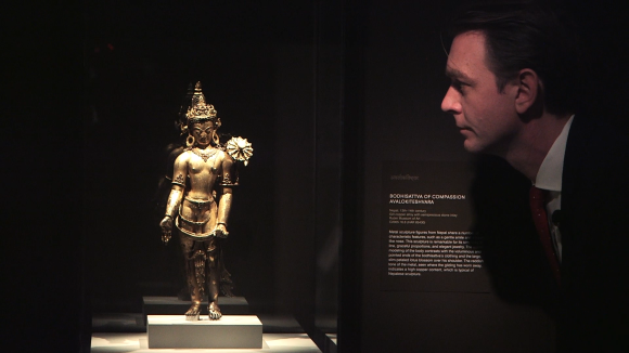 Uomo di profilo e in penombra osserva una scultura buddista, ben illuminata, in una teca
