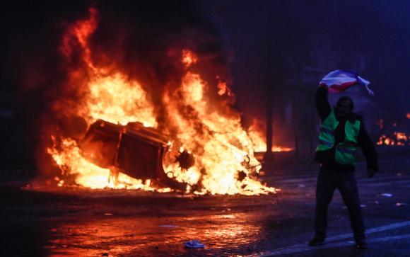 UN uomo con il gilet giallo con la bandiera francese mentre dietro un auto sta bruciando