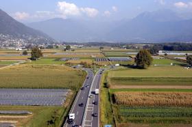 Veduta aerea di un autostrada che scorre in mezzo a campi coltivati; in lontananza, grande rotatoria a doppia corsia