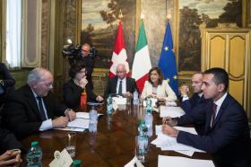 diverse persone sedute attorno a un tavolo con sullo sfondo le bandiere svizzera, italiana ed europea