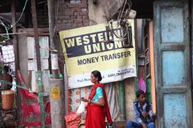 Cartello Western Union appeso tra un pilone e un portone in ambiente urbano indiano; una donna e un bambino davanti