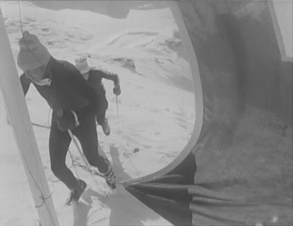 [immagine b/n] Due persone, intravviste dietro una bandiera, scalano una pista innevata con scarponi e racchette da sci