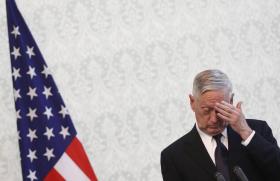 Jim Mattis, al margine dx della foto, guarda in basso e si tocca la fronte; sulla sx, bandiera statunitense