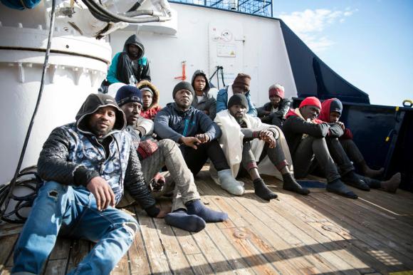 Una decina di uomini di colore, vestiti pesanti e tutti con cuffietta o cappuccio, sul ponte di una nave; guardano in camera