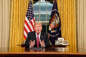 Donald Trump seduto alla scrivania parla gesticolando; dietro, badiera americana e tende/finestra, accanto, foto ritratto