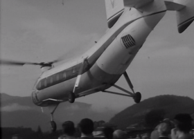 Un elicottero birotore con fusoliera simile a quella di un piccolo aereo si alza in volo, persone sotto; montagne in lontananza