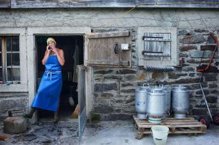 Donna con grembiule plastico fuma una sigaretta sulla soglia dlela porta di una costruzione in pietra; bidoni del latte accanto
