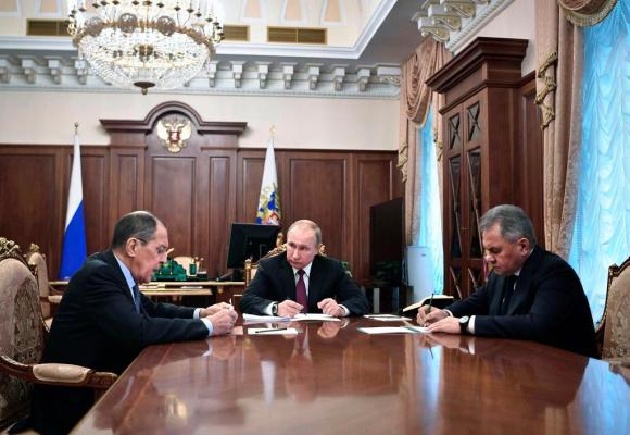 Vladimir Putin seduto a un tavolo tra Lavrov e Shoigu in una sala con le pareti in legno e un grande lampadario