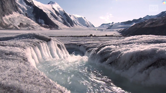 Veduta di un ghiacciaio (non identificato), con montagne innevate ai lati e acqua che scorre in primo piano