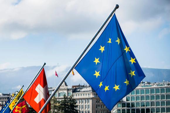 La bandiera euroepa in primo piano e quella svizzera dietro. Sullo sfondo la città di Ginevra