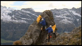 Due alpinisti assicurati lustrano una vetta rocciosa con un lungo telo giallo tenuto ciascuno da un estremità