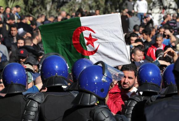 In primo piano poliziotti in tenuta antisommossa, di schiena; di fronte a loro manifestanti con bandiera algerina