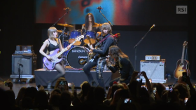 Gruppo rock di 4 persone su un palcoscenico in un momento nel quale bassista, chitarrist e cantante si avvicinano