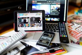 Un laptop aperto, un tablet, un cellulare e due telecomandi appoggiati su un tavolo con giornali; sugli schermi: notizie
